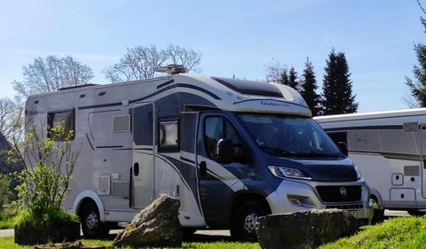 Ein moderner Wohnmobil steht auf einem grünen Feld mit einigen Felsen im Vordergrund. Im Hintergrund sind Bäume und ein klarer Himmel zu sehen.
