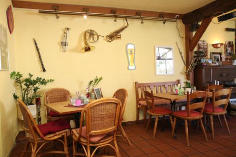 Ein gemütlicher Innenraum mit Tischen und Stühlen aus Rattan. An der Wand hängen Musikinstrumente und ein Bierglas, das eine einladende Atmosphäre schafft.