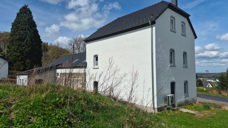 Ein modernes, weißes Haus steht auf einem Hügel in einer ländlichen Umgebung. Im Hintergrund sind Bäume und ein blauer Himmel zu sehen.