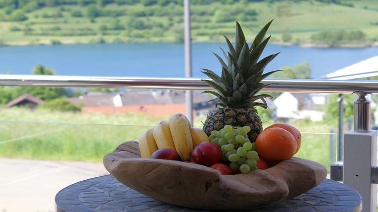 Ein Obstkorb mit Ananas, Bananen, Trauben und einer Orange steht auf einem Tisch. Im Hintergrund ist ein schöner Aussicht auf einen See und grüne Hügel zu sehen.