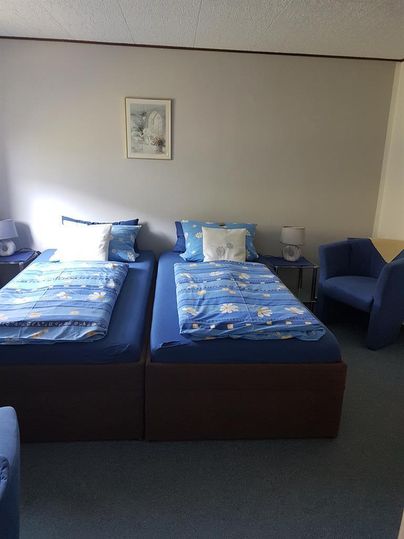 Ein einfach eingerichtetes Zimmer mit zwei einzelnen Betten und blauen Bettdecken. Neben den Betten befinden sich zwei Stühle und ein kleiner Tisch.