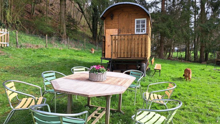 A cozy spot with a colorful table and chairs outside. In the background, there is a wooden house among the trees.