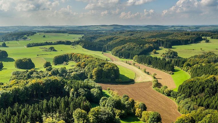 Eine weitläufige Landschaft mit sanften Hügeln, Wäldern und Wiesen. Der Himmel ist blau mit einigen Wolken.