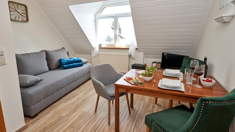 A cozy living area with a gray couch and a small dining table. The room has large windows and a pleasant, inviting atmosphere.