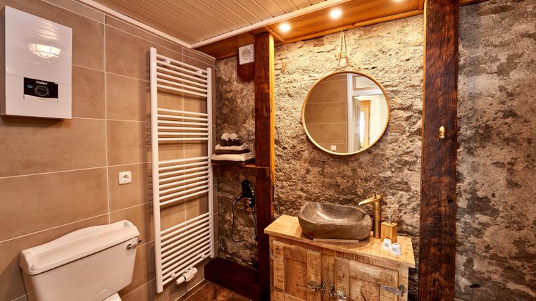 A modern bathroom with a wooden sink and a round mirror. The walls are partially decorated with stone and wood.