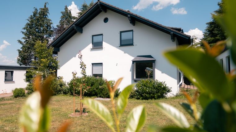 Ein modernes, weißes Haus umgeben von einem gepflegten Garten. Die Sonne scheint und der Himmel ist klar.