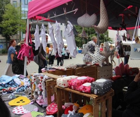 A market stall with handcrafted children's items and colorful stuffed animals. In the background, people can be seen exploring the market.