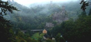 Eine beeindruckende Landschaft mit einer Burg, die von Nebel umgeben ist. Im Vordergrund sind Bäume und einige Gebäude sichtbar.