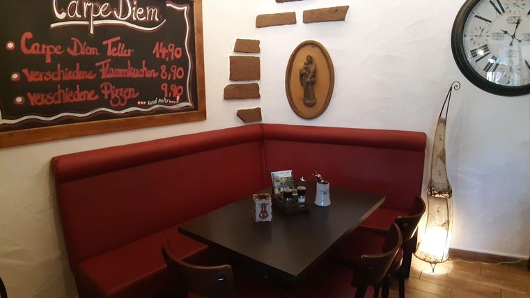 Eine gemütliche Ecke in einem Restaurant mit rotem Sitzpolster und einem Tisch. An der Wand hängt eine Tafel mit dem Menü und eine große Uhr.