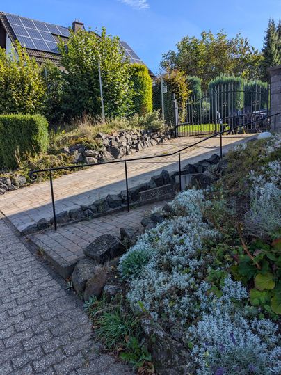 Een geplaveide oprit leidt naar een tuin met struiken en bloemen. Op de achtergrond zijn een hek en een huis met zonnepanelen te zien.