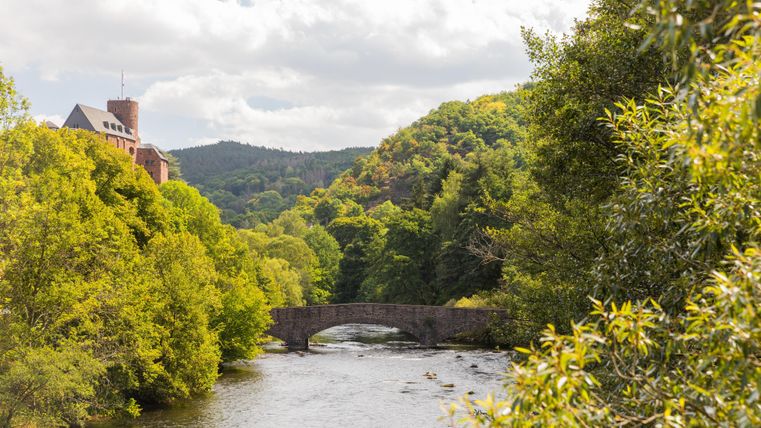 Een schilderachtig landschap met een rivier en een boogbrug. Omringd door groene bomen en zachte heuvels onder een bewolkte lucht.