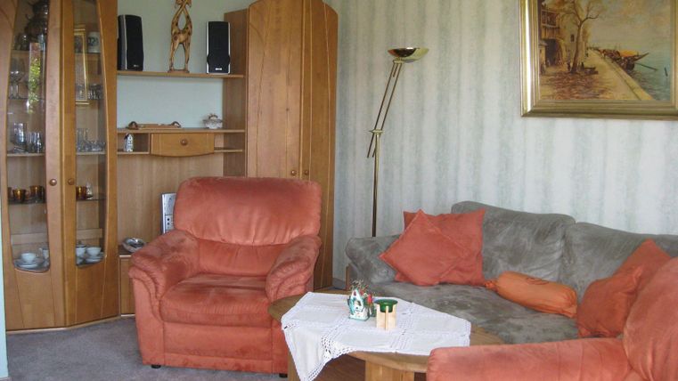 Een gezellige woonkamer met een grijze bank en twee oranje fauteuils. Aan de wand hangt een schilderij, en in de kamer staan houten kasten met decoratieve objecten.