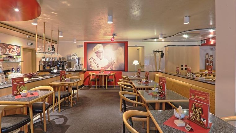 Gemütliches Café mit Holzmöbeln, Tischen und Stühlen. An der Wand ein großes Bild eines Bäckers. Rote Akzente und Menükarten auf den Tischen.