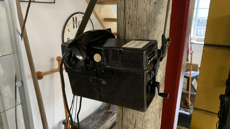 Ein altes Wandtelefon mit Kurbel ist an einem Holzpfosten montiert. Im Hintergrund sind Schilder und ein Treppenaufgang zu sehen.