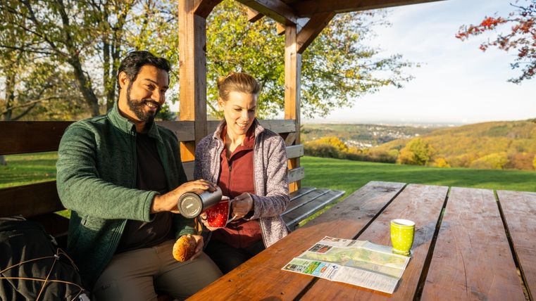 A couple is sitting in a relaxation area under a wooden pavilion, enjoying a warm drink. In the background, there are trees and a picturesque landscape.