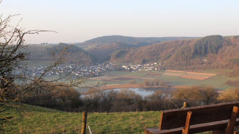 A picturesque landscape with gentle hills and a small village in the valley. In the foreground, there is a wooden bench that offers a beautiful view of the surroundings.