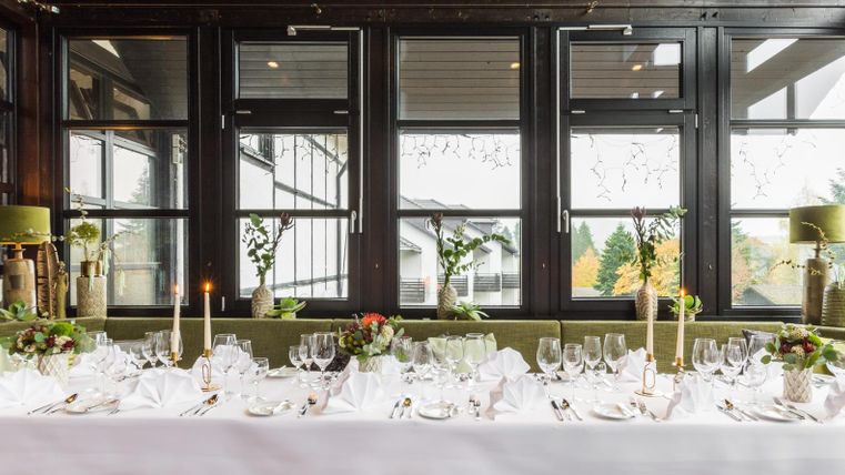 Ein elegant gedeckter Tisch in einem modernen Restaurant mit großen Fenstern. Die Dekoration umfasst Kerzen und Blumenelemente.