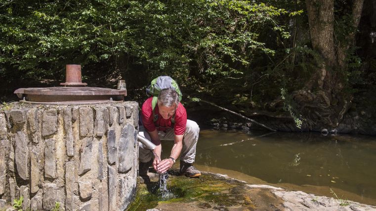 A man is leaning over a well and drawing water. In the background, there are trees and a calm river.
