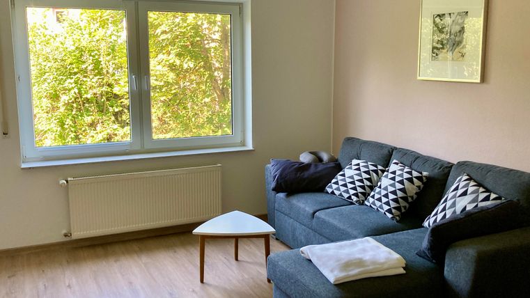 Ein gemütliches Wohnzimmer mit einem grauen Sofa und dekorativen Kissen. Große Fenster bieten einen Blick auf die grüne Umgebung.