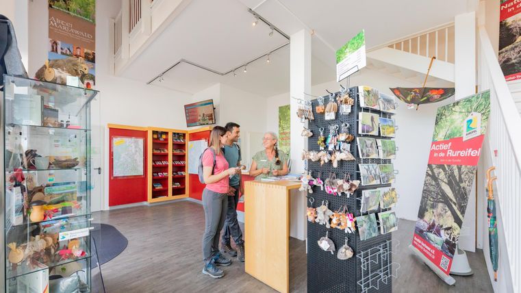 Een ontvangstgebied met een verkooppresentatie van souvenirs. Twee bezoekers staan aan de receptie en praten met een medewerker.