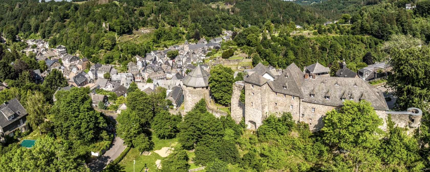 Panoramablick auf Monschau mit der Burg im Vordergrund, umgeben von grüner Landschaft und bewaldeten Hügeln unter blauem Himmel mit Wolken., © Eifel Tourismus GmbH, Dominik Ketz