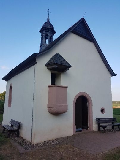 Eine kleine, charmante Kapelle mit einem glockenförmigen Türmchen und einer hellen Fassade. Bänke laden zum Verweilen im Freien ein.
