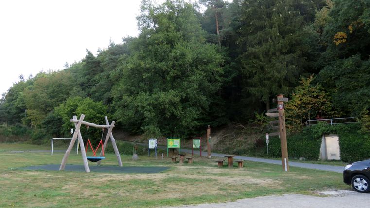 Een speeltuin met een schommel en enkele zitplaatsen in een groen gebied. Op de achtergrond zijn bomen en een pad te zien.