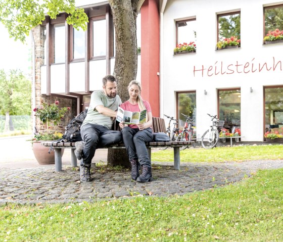 Zwei Personen sitzen auf einer Bank vor dem Heilsteinhaus in Einruhr, lesen eine Broschüre. Fahrräder stehen vor dem Gebäude, das mit Blumen geschmückt ist., © -@Eifel Tourismus GmbH, AR-shapefruit-AG