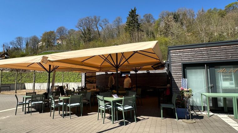 A cozy outdoor area of a restaurant with green tables and chairs. In the background, there are umbrellas and a green landscape visible.