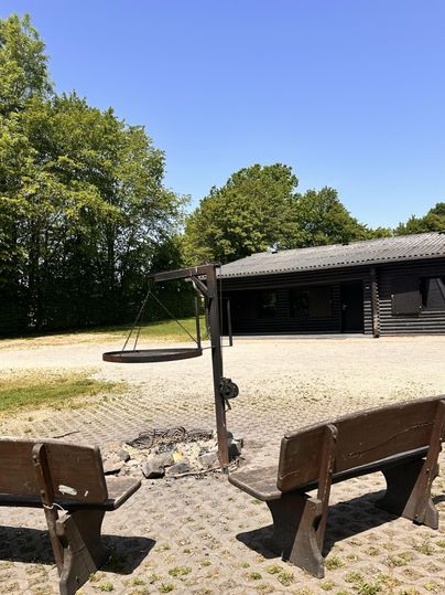 Ein ruhiger Platz im Freien mit zwei Bänken und einem Lagerfeuerbereich. Im Hintergrund steht ein Holzgebäude und die Sonne scheint.