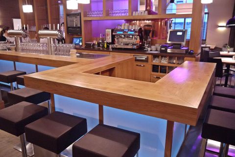 Eine moderne Bar mit einem Holztheke und stilvollen Hockern. Der Bereich ist gut beleuchtet und hat eine einladende Atmosphäre.