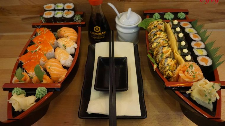 Ein ansprechendes Sushi-Set auf zwei Booten, dekoriert mit verschiedenen Sushi-Sorten und Beilagen. Ein Sojasauce-Behälter und Essstäbchen vervollständigen das Bild.