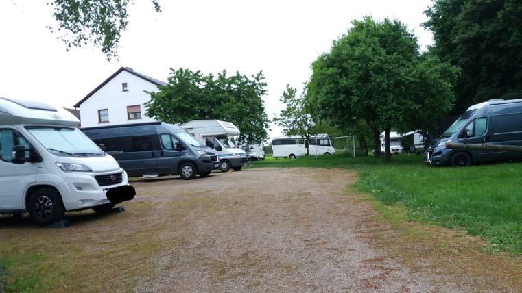 Wohnmobilstellplatz mit mehreren geparkten Wohnmobilen, umgeben von Bäumen und einem weißen Gebäude im Hintergrund.
