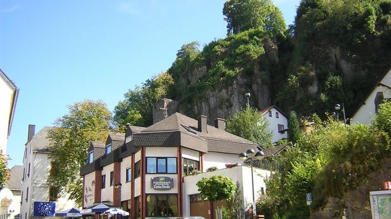 Pizzeria Costa Verde in front of a rock wall, surrounded by trees. Several cars are parked in front of it, sun umbrellas are ready. Blue sky in the background.