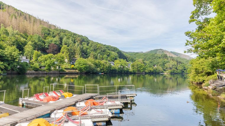 Ein ruhiger See mit einem Steg und bunten Ruderbooten. Umgeben von grünen Bäumen und sanften Hügeln.