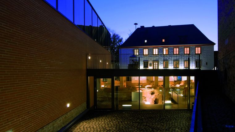 Eine moderne Architektur mit einem Glasgebäude und einem historischen Haus im Hintergrund. Es ist Abend und der Himmel hat einen schönen Blau-Ton.