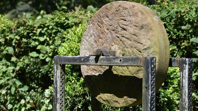 Ein großes, runde Stein auf einem Ständer, umgeben von grünen Sträuchern. Die Oberfläche des Steins zeigt alte Gebrauchsspuren.