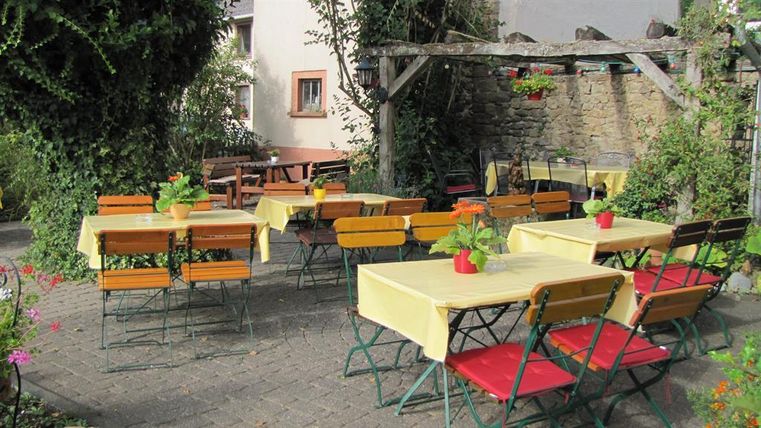 Ein gemütlicher Außenbereich mit mehreren Tischen und Stühlen. Die Tische sind mit gelben Tischtüchern und kleinen Pflanzen dekoriert, umgeben von einer grünen, blühenden Landschaft.
