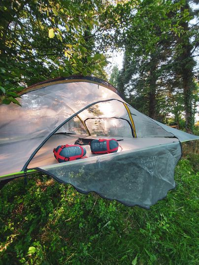Een campingtent op een weiland, omringd door bomen. Twee rugzakken liggen op het tentgrond.