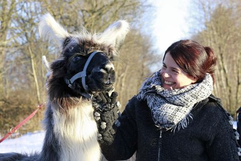 A woman smiles while petting a lamb. In the background, trees and snow can be seen.