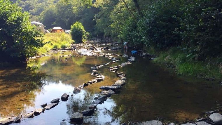 Een rustige rivier stroomt door een bosrijk gebied, met platte stenen die in het water liggen. Op de achtergrond zijn bomen en enkele tenten zichtbaar.