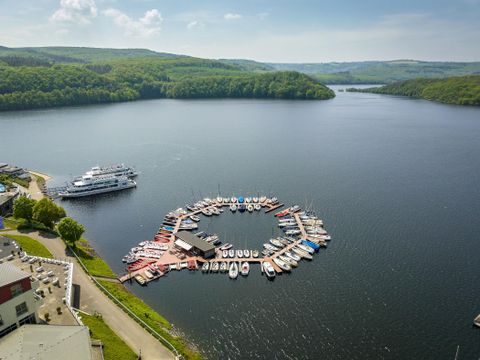 An idyllic landscape with a picturesque harbor and many boats on a tranquil lake. In the background, green hills and a clear sky can be seen.