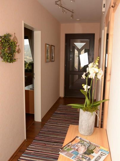 A bright hallway with a flower plant on a table. Pictures hang on the walls and the floor is covered with a striped rug.