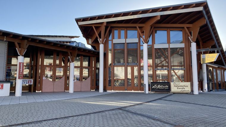 Ein modernes Gebäude mit großen Fenstern und Holzverkleidung. Davor befindet sich ein gepflasterter Platz.