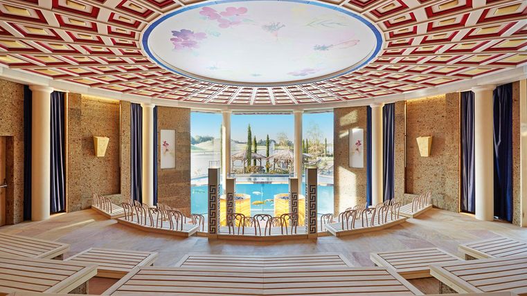 Een elegante zaal met een kunstzinnig versierde plafond en houten banken. Grote ramen bieden uitzicht op een prachtig landschap.