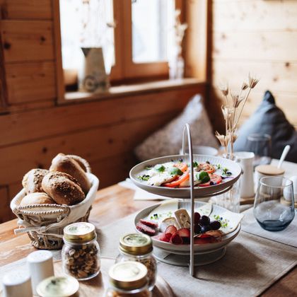 Ein ansprechend gedeckter Tisch aus Holz mit verschiedenen Frühstücksoptionen. Darauf sind frische Brötchen, Obst und Gläser mit Müsli und Marmelade zu sehen.