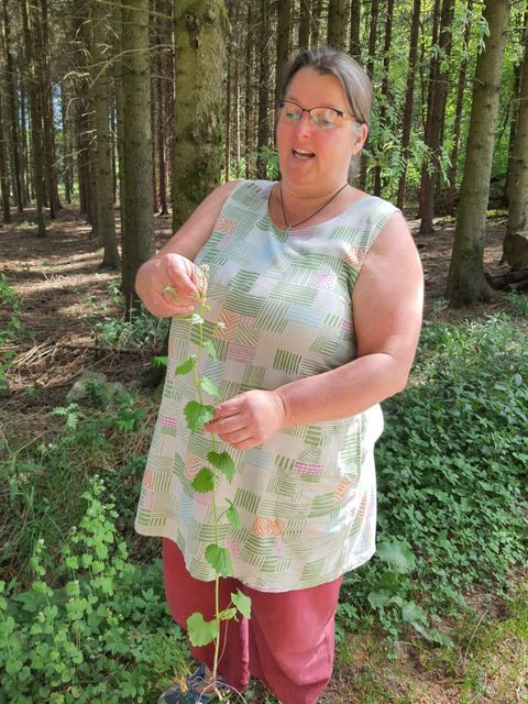 Een vrouw staat in het bos en houdt een plantendraad in haar hand. Ze draagt een lichte top en heeft lang haar.