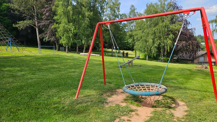 Eine Schaukel in einem grünen Park mit Bäumen im Hintergrund. Der Himmel ist blau und die Wiese ist gut gepflegt.