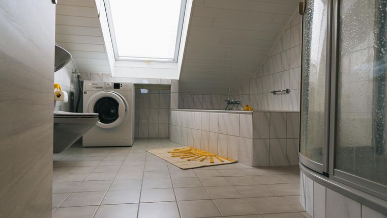 Een moderne badkamer met een dakraam, een wasmachine en een douche. De ruimte is licht en helder betegeld, met een geel tapijt op de vloer.