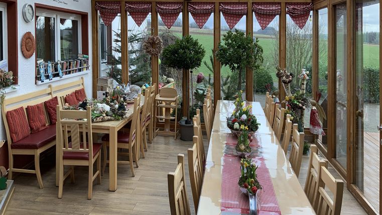 A bright winter garden with long tables and colorful chairs. Flower arrangements and a green view create an inviting atmosphere.